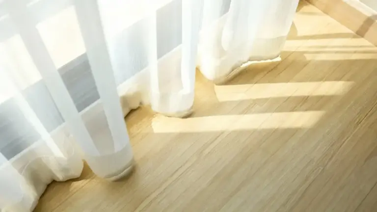Tende bianche leggere che sfiorano un pavimento in legno chiaro illuminate dal sole